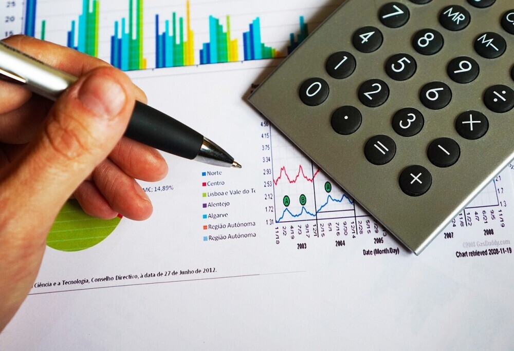 image of fingers holding a pen with a calculator and graph sheet. This image related to financial literacy in the real estate business.